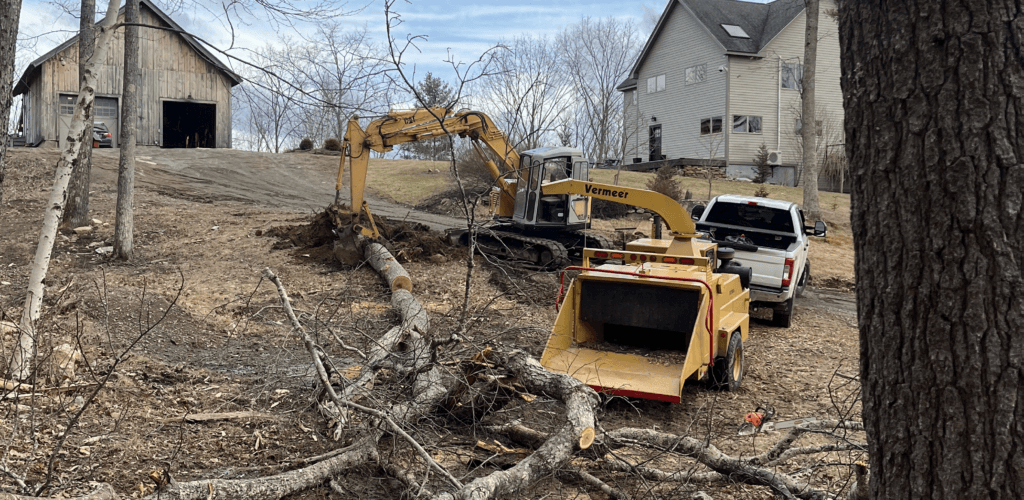 Excavator and felled logs beside a house and barn during a tree removal